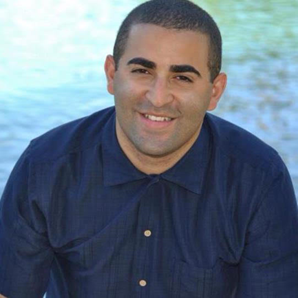 Tactile Description: when first introducing, hands got nervous and exciting at same time, feeling curly hairs on the arms, introducing my curt hairs on arm as my tactile name. Image Description: Dominican male wearing a blue buttoned down collared shirt. The image zoomed in to his head and shoulders. He sits in front of a body of water, smiles directly at the camera.