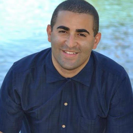 Tactile Description: when first introducing, hands got nervous and exciting at same time, feeling curly hairs on the arms, introducing my curt hairs on arm as my tactile name. Image Description: Dominican male wearing a blue buttoned down collared shirt. The image zoomed in to his head and shoulders. He sits in front of a body of water, smiles directly at the camera. 