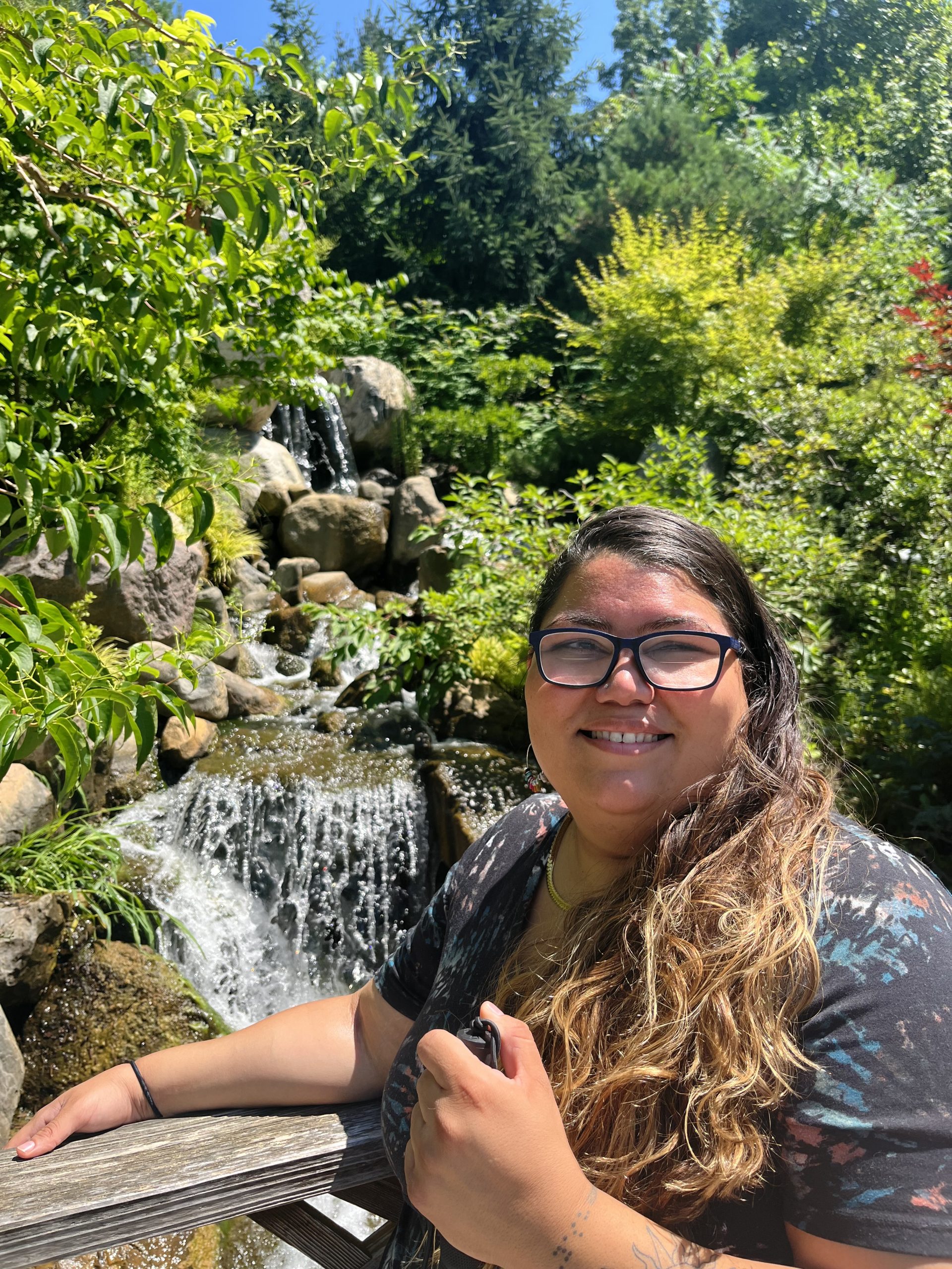 Tactile Description: A strong-willed fashionista who hugs with hearts drawn on back and kisses on check, easily spreading a bubbly sensation with a hint of infectious giggles. Image Description: Yash is smiling at the camera and is standing in front of a waterfall, surrounded by trees, rocks, and bushes. 