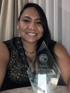 A woman with dark brown hair, dark brown eyes, and tan skin. She is wearing a black floral lace shirt with a necklace and is smiling in front of an award with her name on it.